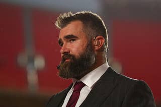 A man with a beard and wearing a suit and tie stands on a red carpet, wearing a headset microphone, in a well-lit venue