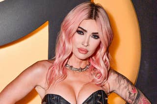 Person with pink hair in a leather studded outfit poses confidently; has tattoos and wears a silver necklace