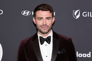 Person in a velvet suit and bow tie at a formal event, in front of a backdrop with sponsor logos