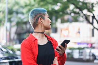 Person with short, brightly dyed hair, wearing a black top, red jacket, and choker necklace, looking to the side while holding a smartphone