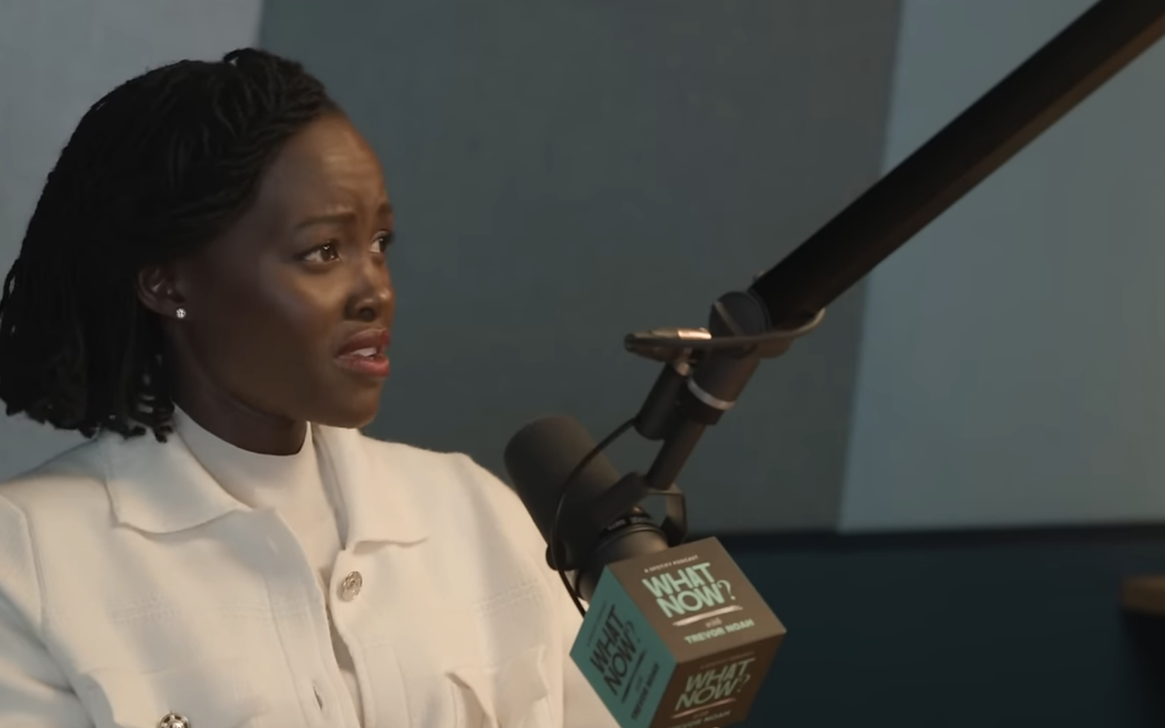 Lupita Nyong'o speaks into a microphone during the "What Now?" podcast with Trevor Noah, discussing African diaspora topics