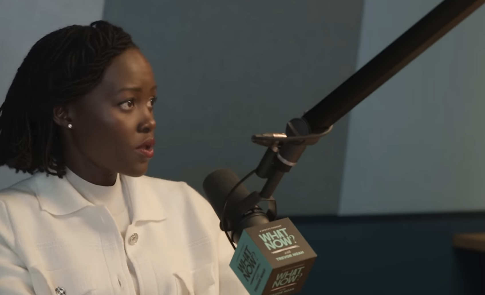 Lupita Nyong'o in a white outfit talks into a microphone on Trevor Noah's podcast, "What Now?" displayed on a YouTube video page